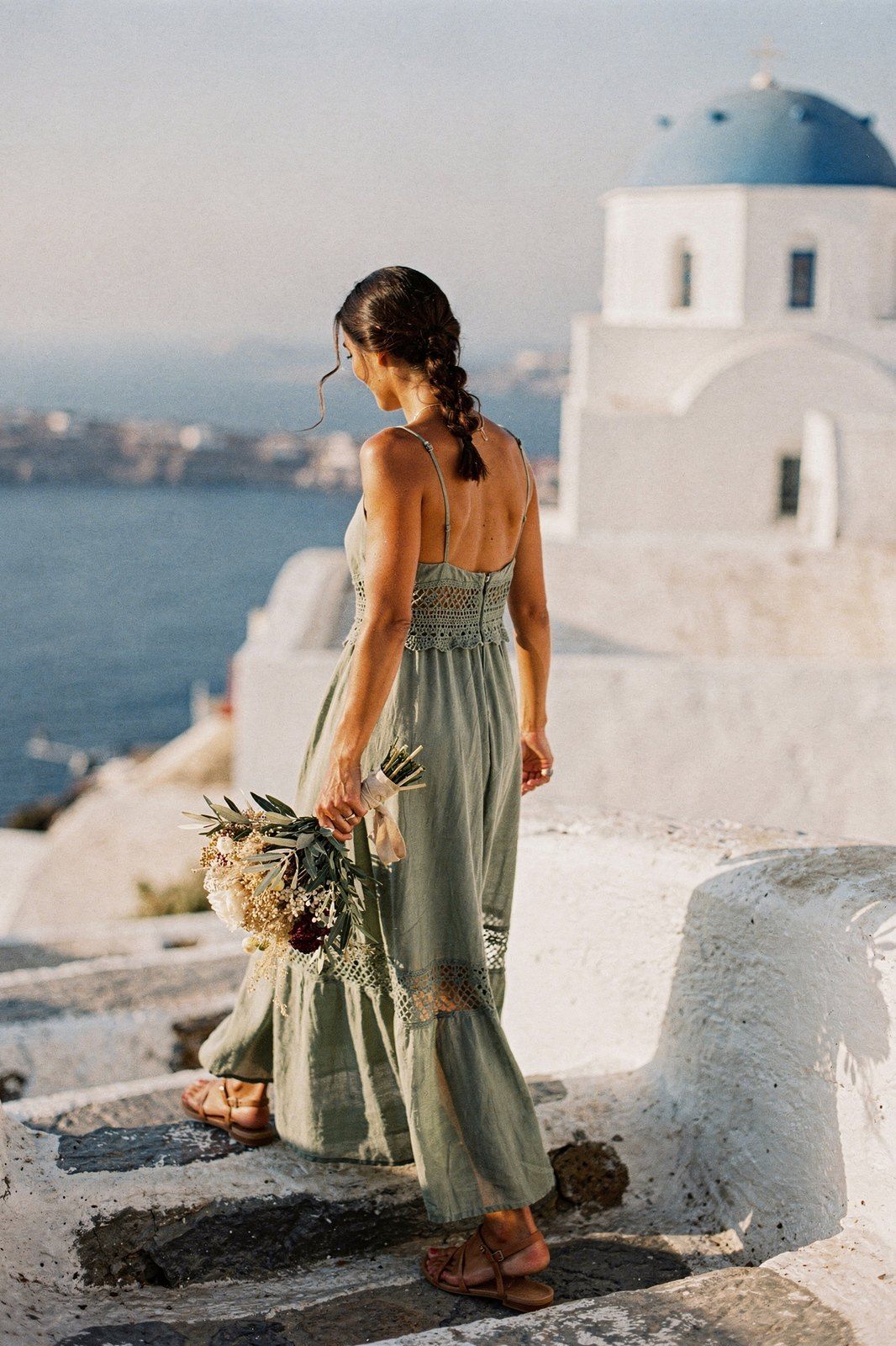 Κουμπάρα με boho φόρεμα σε φασκομηλί και πλεξούδα σε σκαλιά νησιού, μπροστά σε ξωκλήσι με μπλε τρούλο